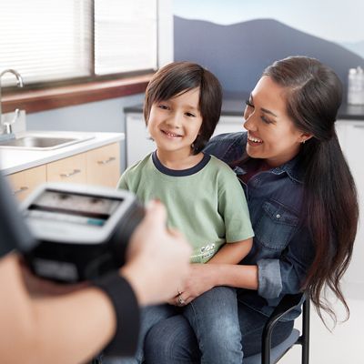 Un profesional de la salud sostiene el dispositivo de valoración de la visión Spot® (visible parcialmente en primer plano), y un paciente joven con su madre están sentados en plano medio