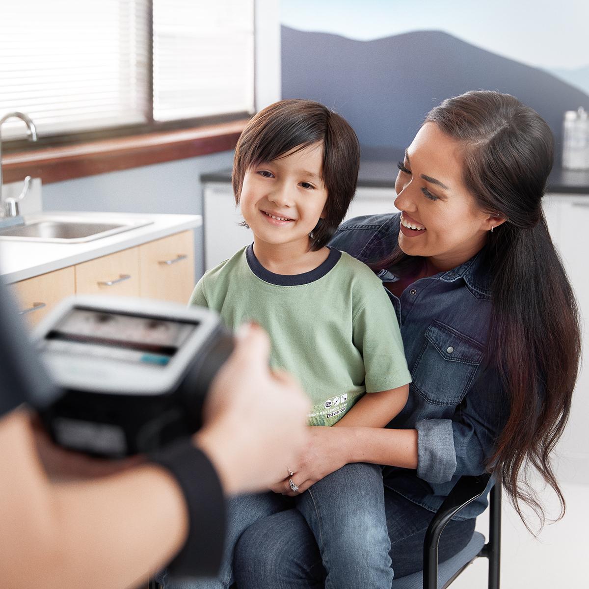 Welch Allyn Spot Vision Screener that quickly detects amblyopia vision issues in pediatric patients being held by clinician (partially seen in foreground), and young patient and mother, seated, in mid-ground