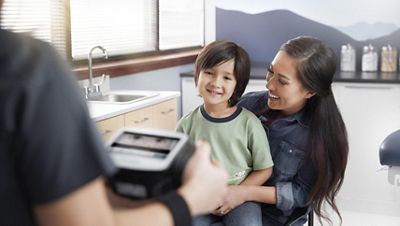 A smiling child sits on a woman's lap as a clinician in the foreground holds a vision screener.