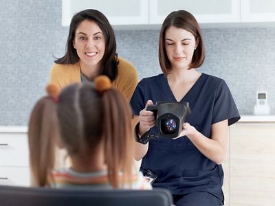 Clinician uses the Welch Allyn® Spot® Vision Screener device to screen child’s eye from a 3 foot distance. Mother of the child stands with the clinician, smiling at the child.