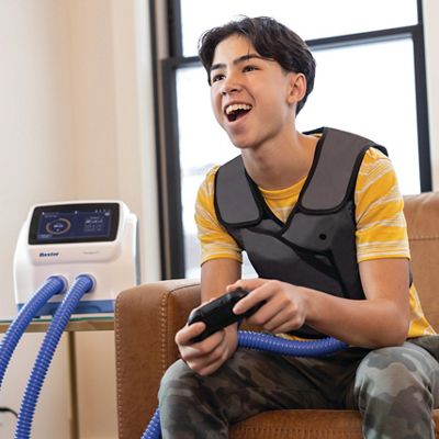 : Image of a teenage boy wearing the vest garment for treatment while playing video games, with the control unit on a table next to him