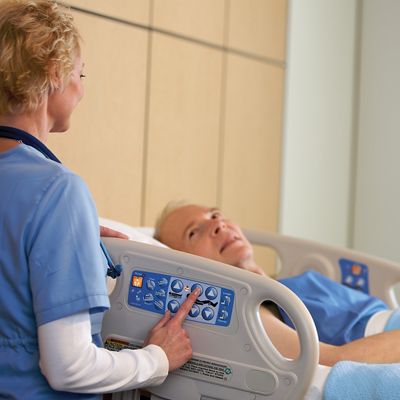 An older male patient lies in a VersaCare Med Surg Bed. A female clinician stands at the bedside, back to camera, using the controls.