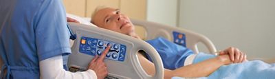 An older male patient lies in a VersaCare Med Surg Bed. A female clinician stands at the bedside, back to camera, using the controls.