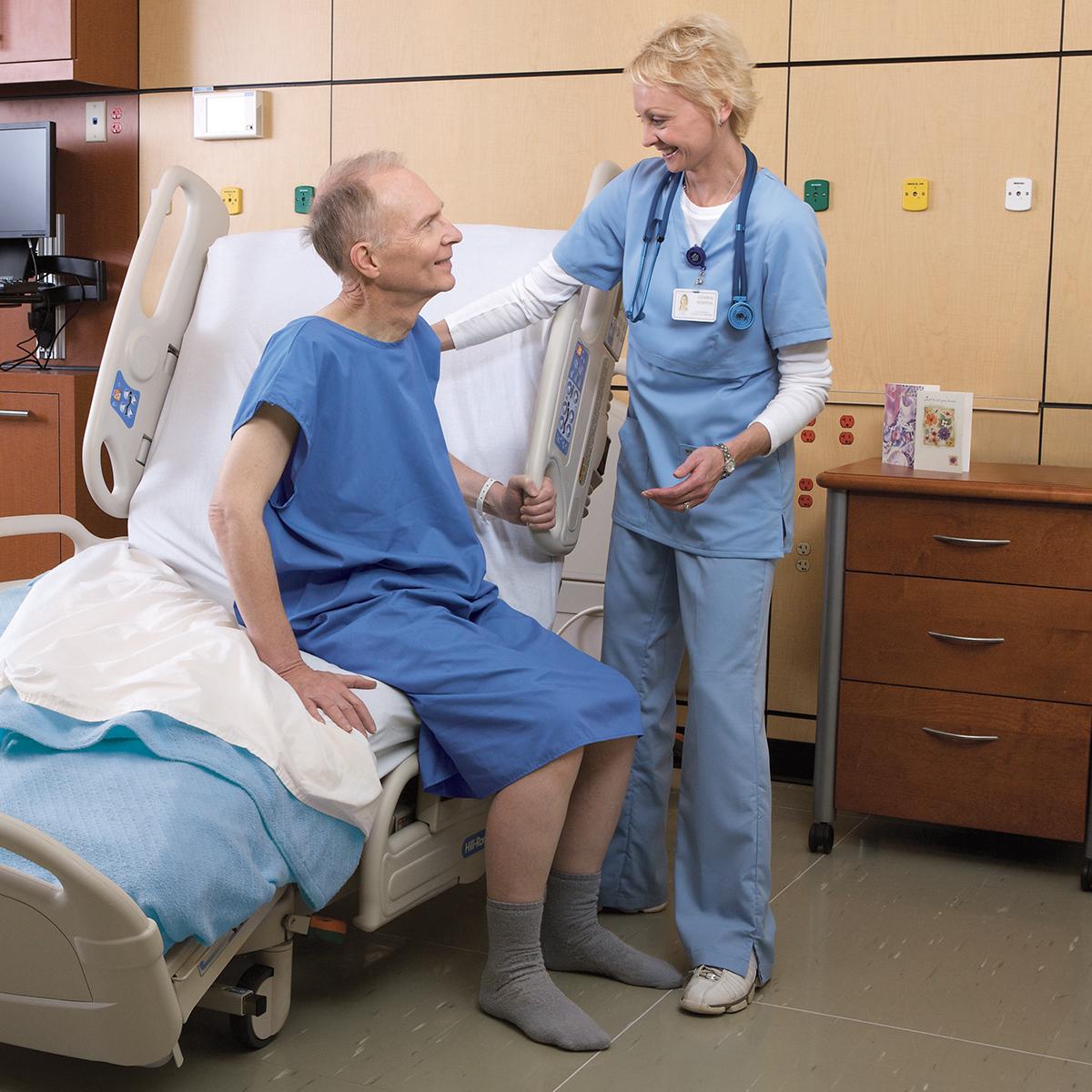 An older male patient sits up on the VersaCare Med Surg Bed. A female clinician stands at the bedside, assisting him with her hand on his shoulder.