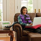 Sistema The&nbsp;Vest, modelo&nbsp;105, chaleco color púrpura usado por una mujer joven sentada en el sofá de la sala de estar con una computadora portátil mientras recibe tratamiento