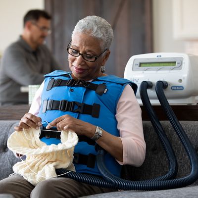 Een oudere vrouw breit in haar woonkamer terwijl ze het The Vest-systeem met een Coastal Blue-vest gebruikt.