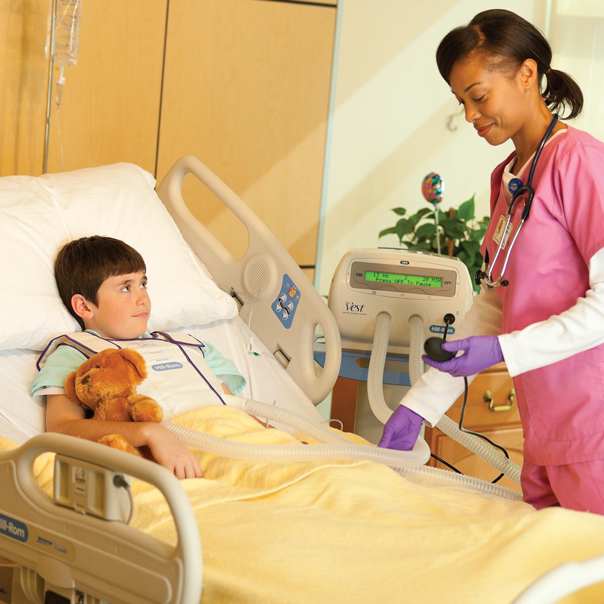 Um menino em uma cama de hospital segura um ursinho de pelúcia enquanto recebe tratamento do sistema Vest, modelo 205.