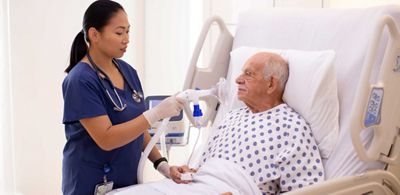 A patient wearing a nasal cannula sits upright in a Hillrom bed while his clinician listens to his heart with a stethoscope