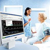 앞쪽 데스크톱 컴퓨터 화면에 Connex 바이탈 관리 소프트웨어가 보이고 뒤에서는 의료진이 환자와 대화하고 있습니다.