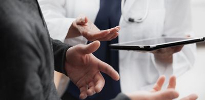 Un médecin montre des données sur une tablette à son patient. Les mains sont représentées dans une conversation active.