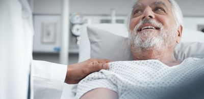 A physician comforts and older male patient, lying in a hospital bed, placing a hand on his shoulder.