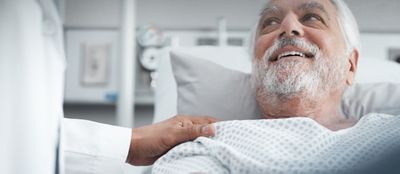 A physician comforts and older male patient, lying in a hospital bed, placing a hand on his shoulder.