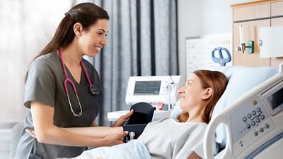 Female nurse puts a blood pressure cuff on a female patient who's in a hospital bed with the Connex Spot Monitor in the background 