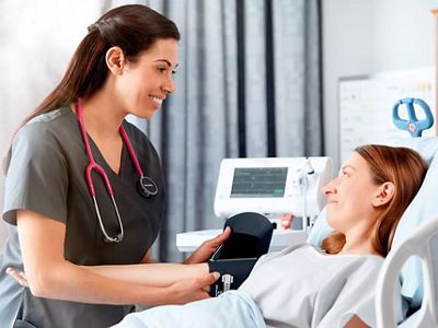 A patient lies in a hospital bed while a clinician measures her blood pressure using the Connex Spot Monitor