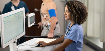 A patient wearing a nasal cannula sits upright in a Hillrom bed while his clinician listens to his heart with a stethoscope