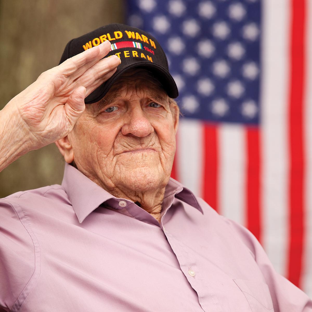 An elderly man wearing a World War II Veteran hat salutes in front of an American flag.