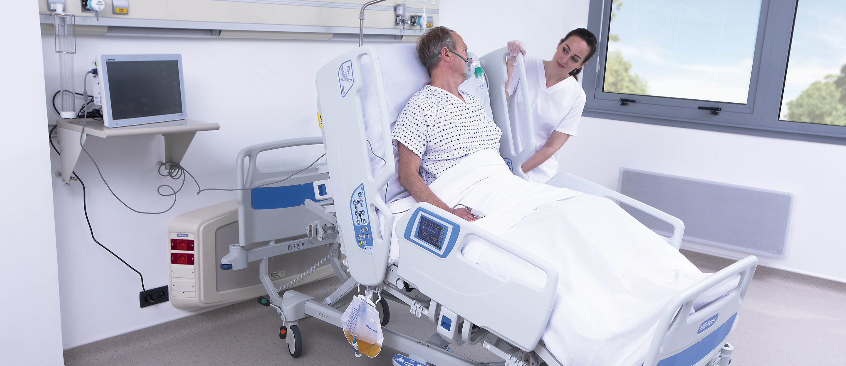 Een pati&euml;nt glimlacht in een Hillrom™ 900 Accella™ Smart Bed. Haar arts staat vlakbij de grafische interface voor de zorgverlener (GCI).