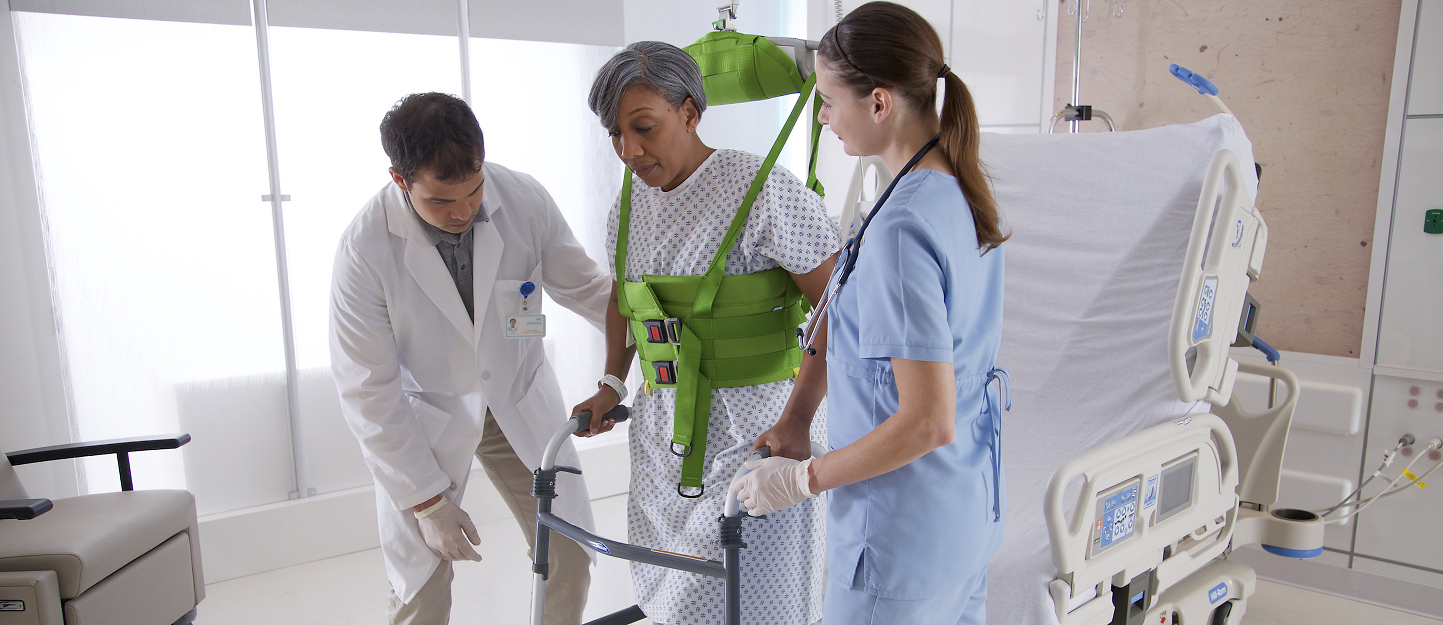 Clinicians help a patient stand and walk using a walker and a Hillrom overhead lift 