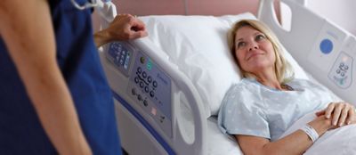 A patient smiles in a Centrella Smart+ Bed. Her clinician stands near the Graphical Caregiver Interface (GCI) Touchscreen.