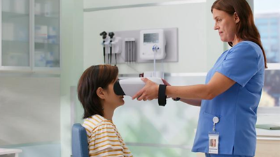 A smiling child sits on a woman's lap as a clinician in the foreground holds a vision screener.