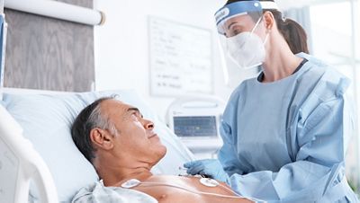 Adult female caregiver standing next to male patient with ECG electrodes showing and connected to The Welch Allyn® Connex®Vital Signs device in right corner.