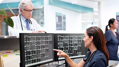 Clinicians review patient data on dual monitors at a central nurses station.